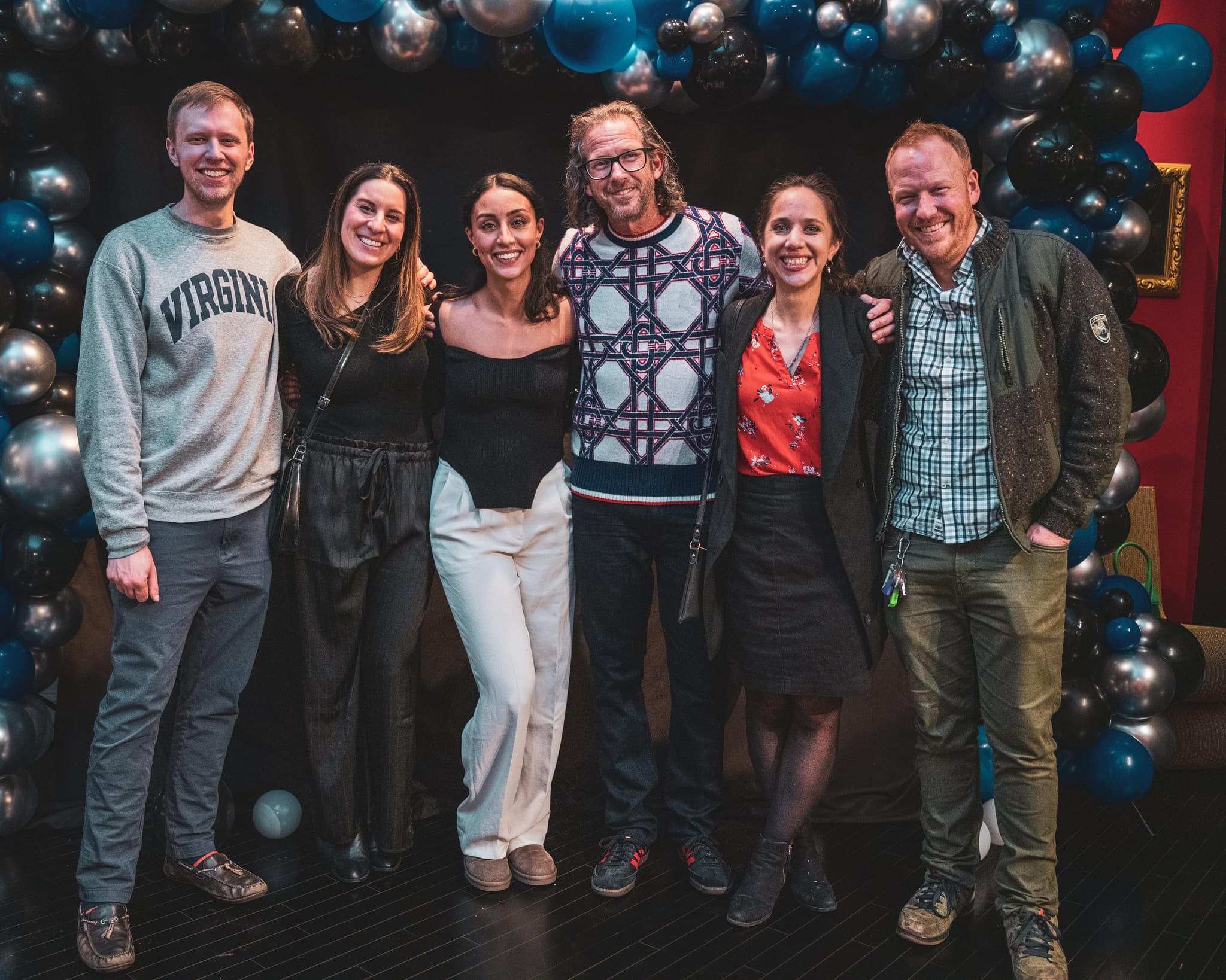 Six friends posing together and smiling in front of a dramatic black and silver balloon arch at the birthday celebration
