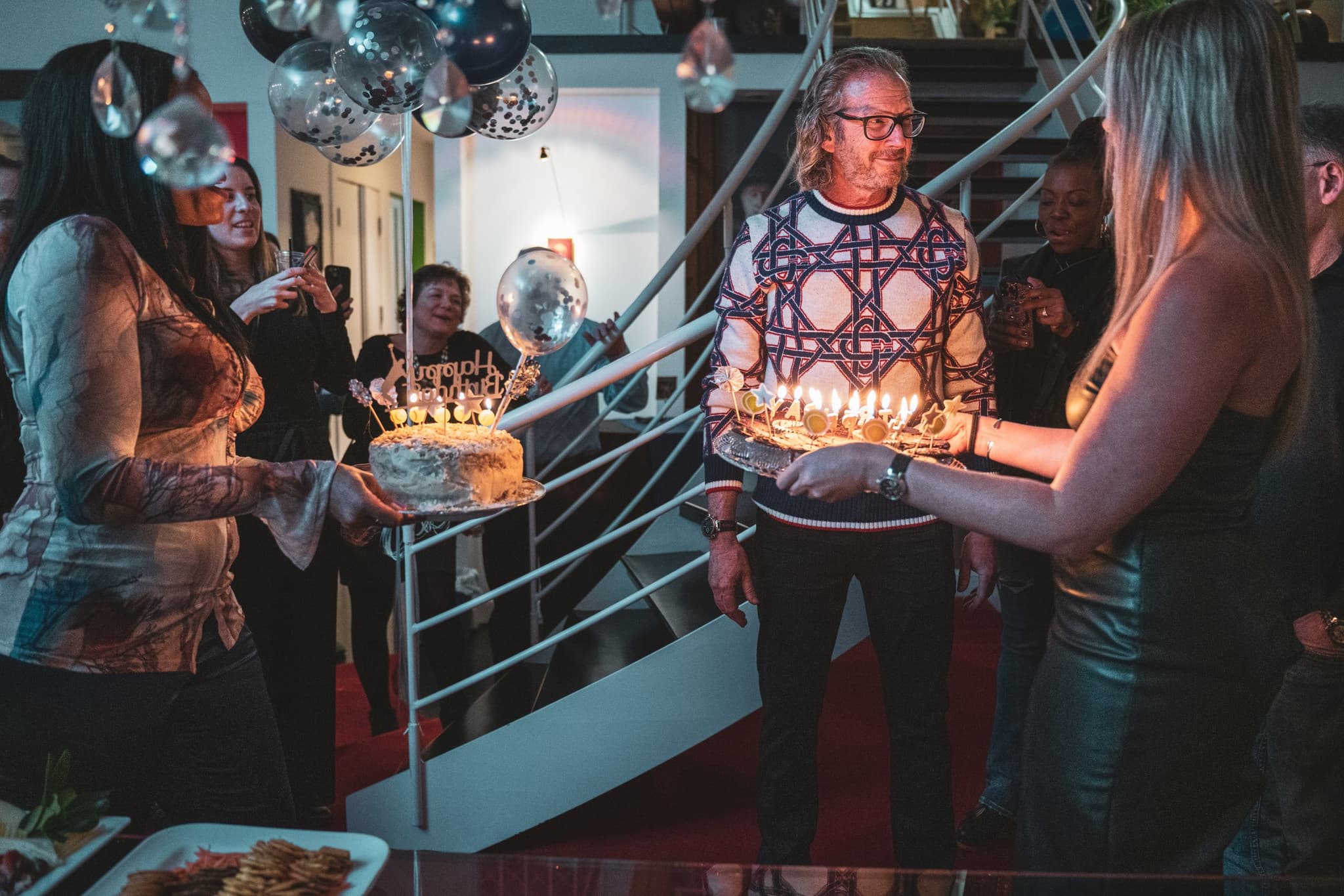 The birthday honoree surrounded by guests as two women present him with a lit birthday cake on a staircase landing