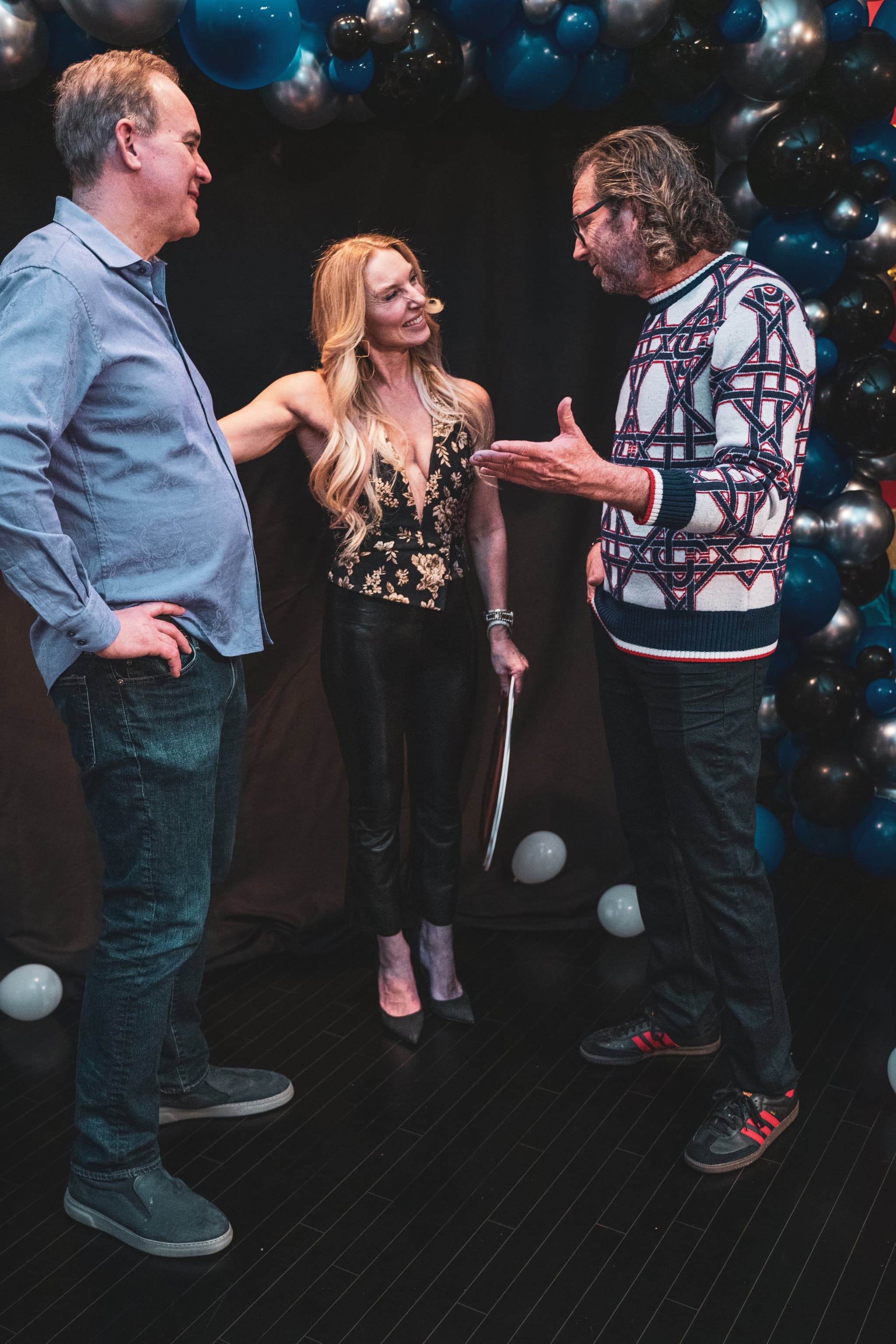 Three guests laugh and chat together beneath a festive black and blue balloon arch at the party