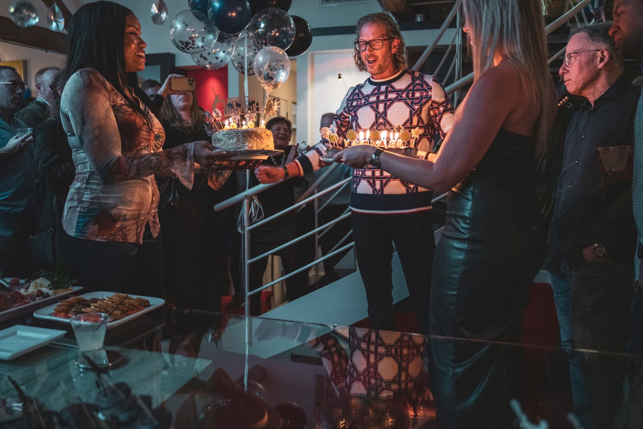 The birthday honoree beams as a lit birthday cake is presented to him amid a crowd of celebrating guests