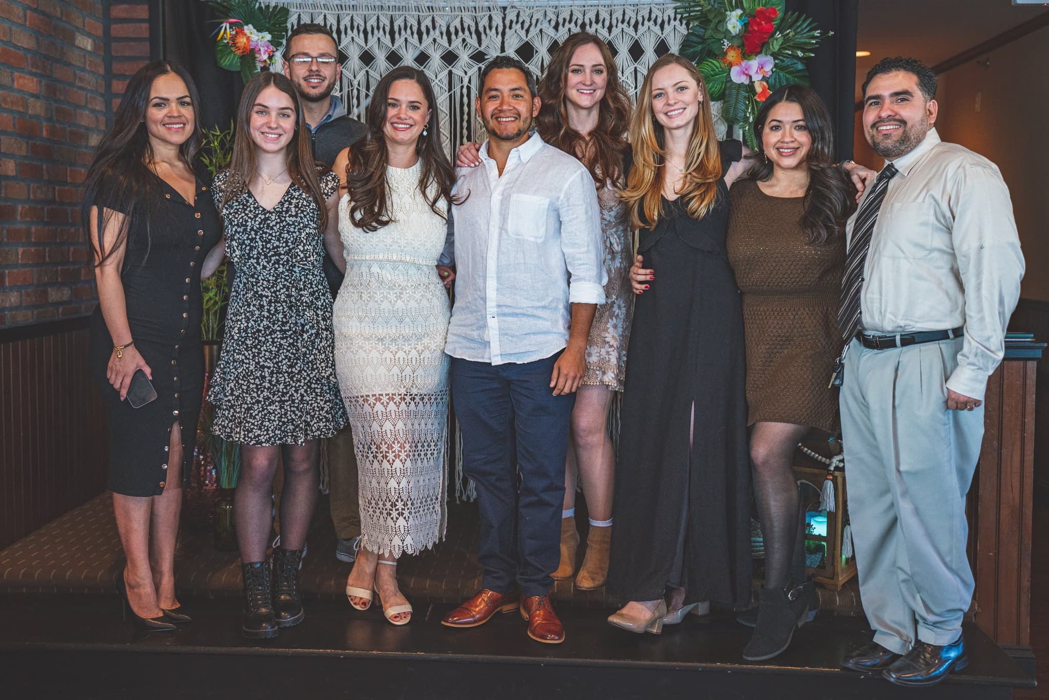 A large group of family and friends posing together in front of a decorative macrame wall hanging adorned with tropical flowers