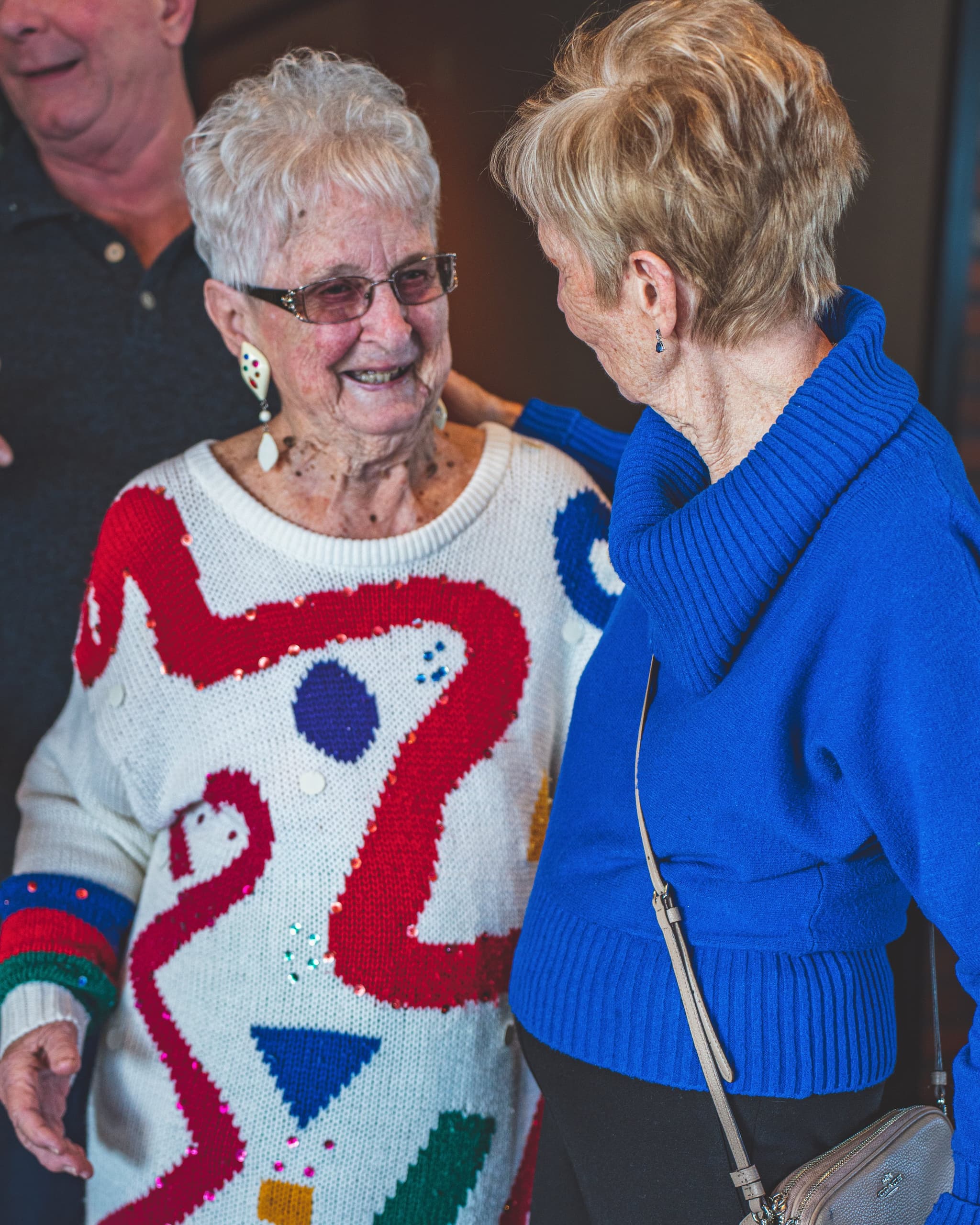 Two elderly women in colorful sweaters sharing a warm, candid conversation at the bridal shower