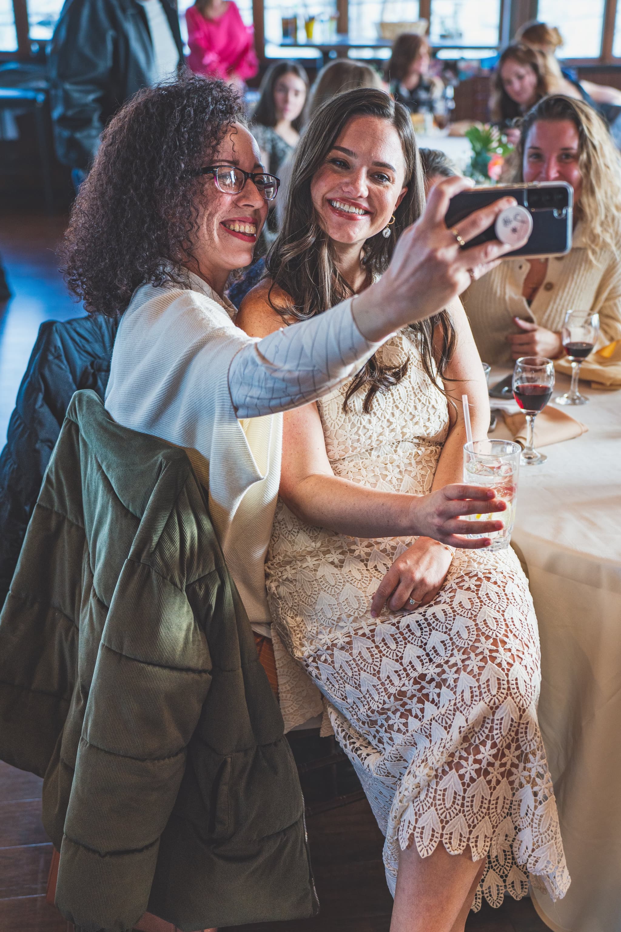 The bride-to-be taking a joyful selfie with a guest while holding a cocktail at a long banquet table