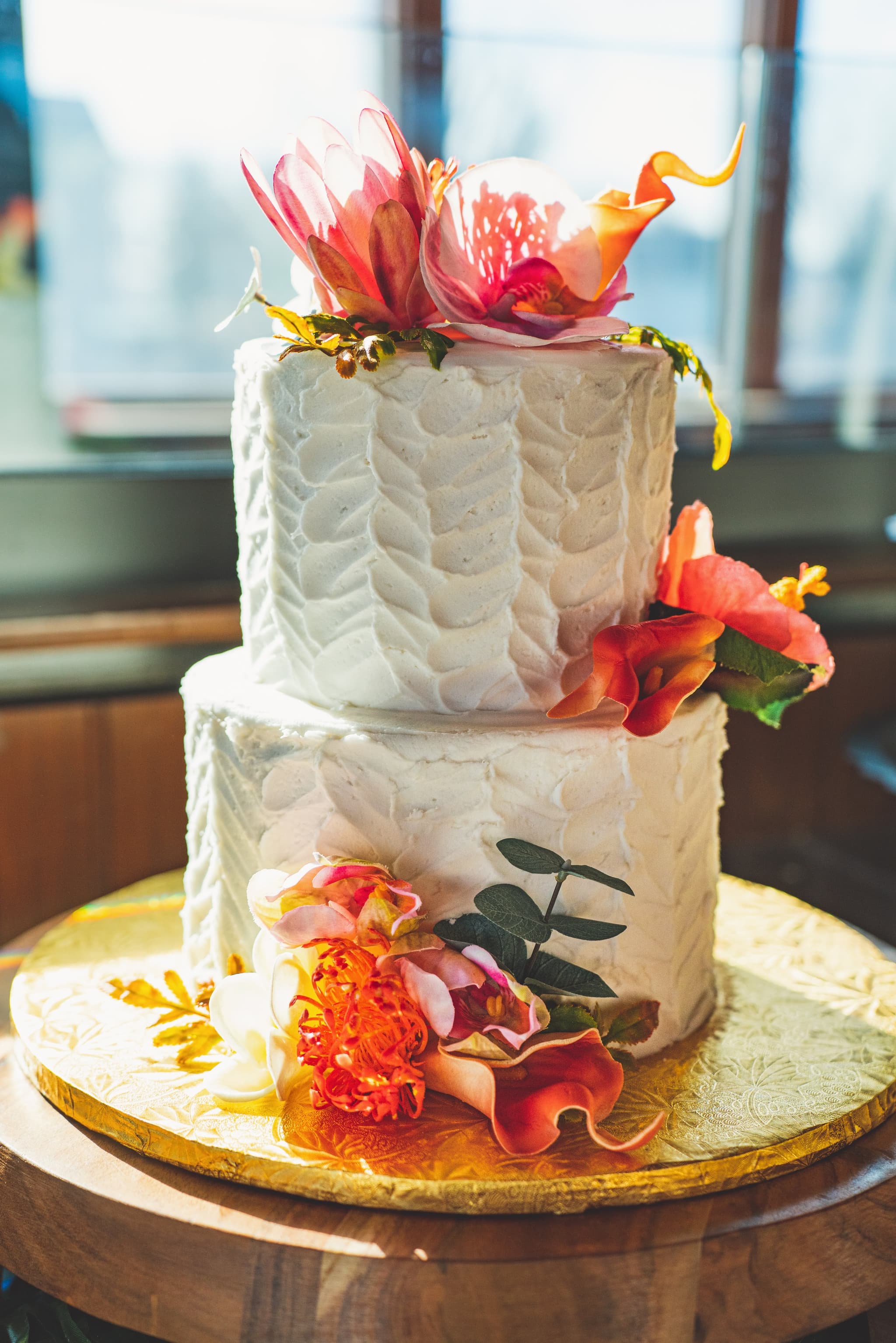 A two-tiered white textured bridal shower cake decorated with vibrant tropical flowers on a wooden cake stand