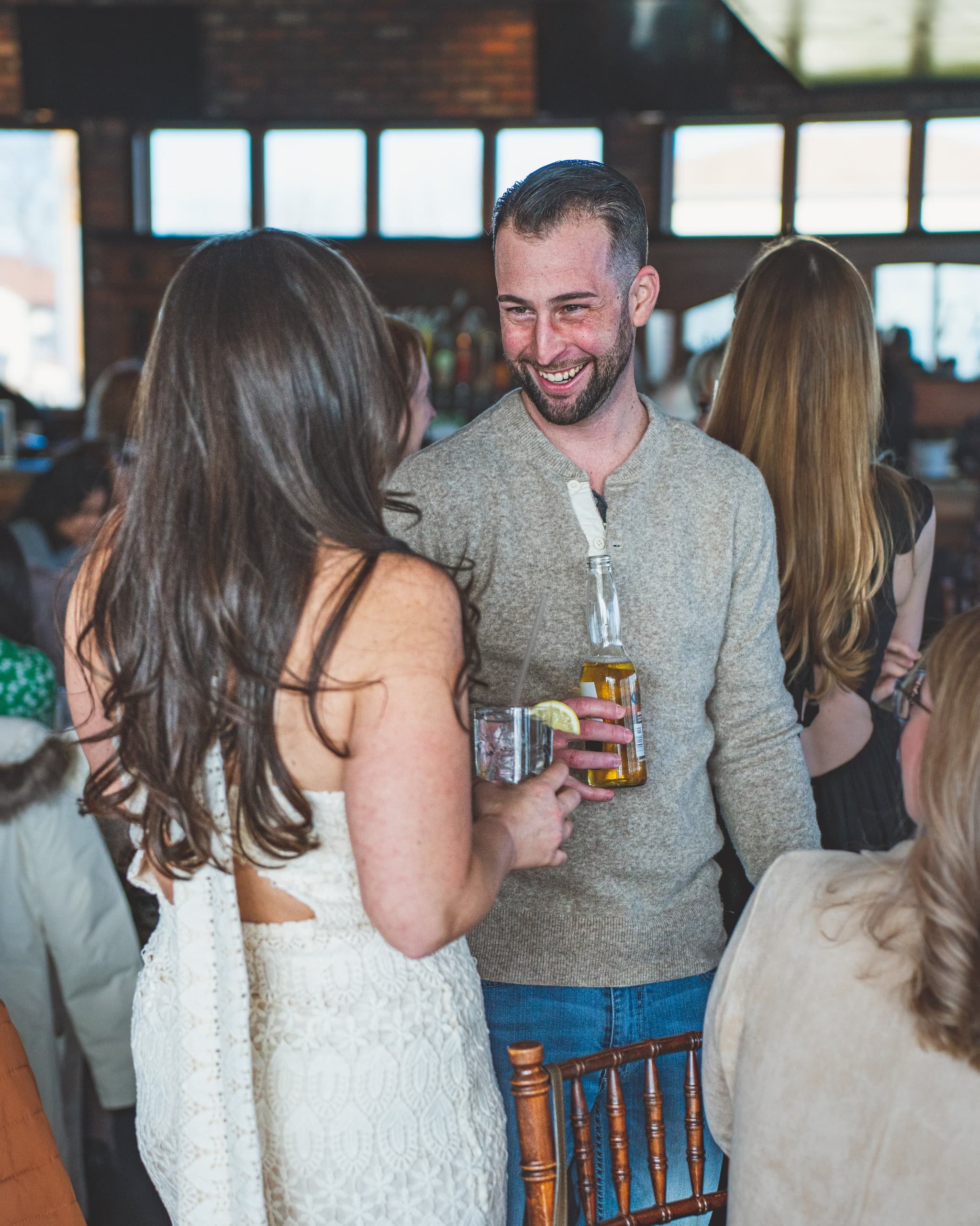 The bride in a white lace dress sharing a candid, laughing conversation with a guest holding drinks at the bridal shower
