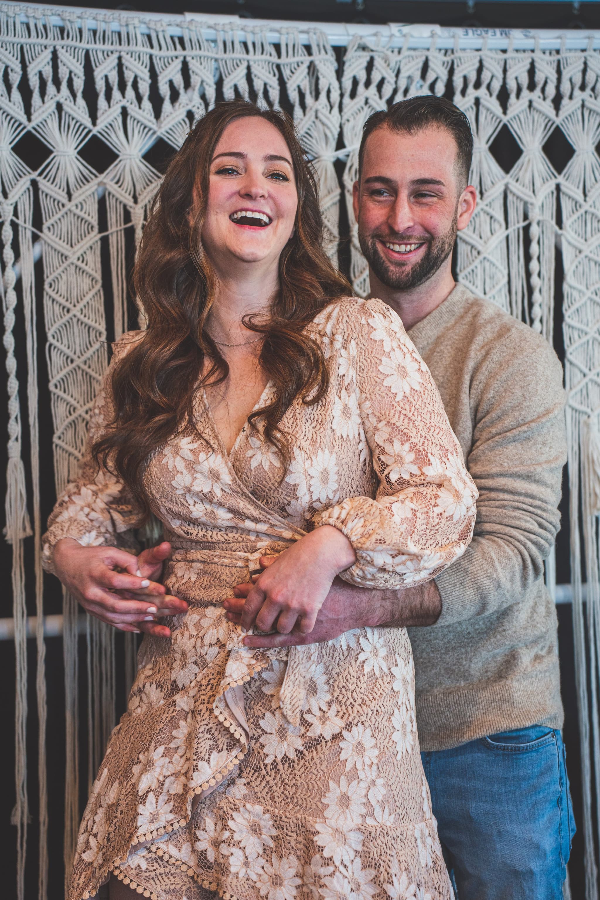 A joyful couple laughing together in front of a boho macrame backdrop at the bridal shower celebration