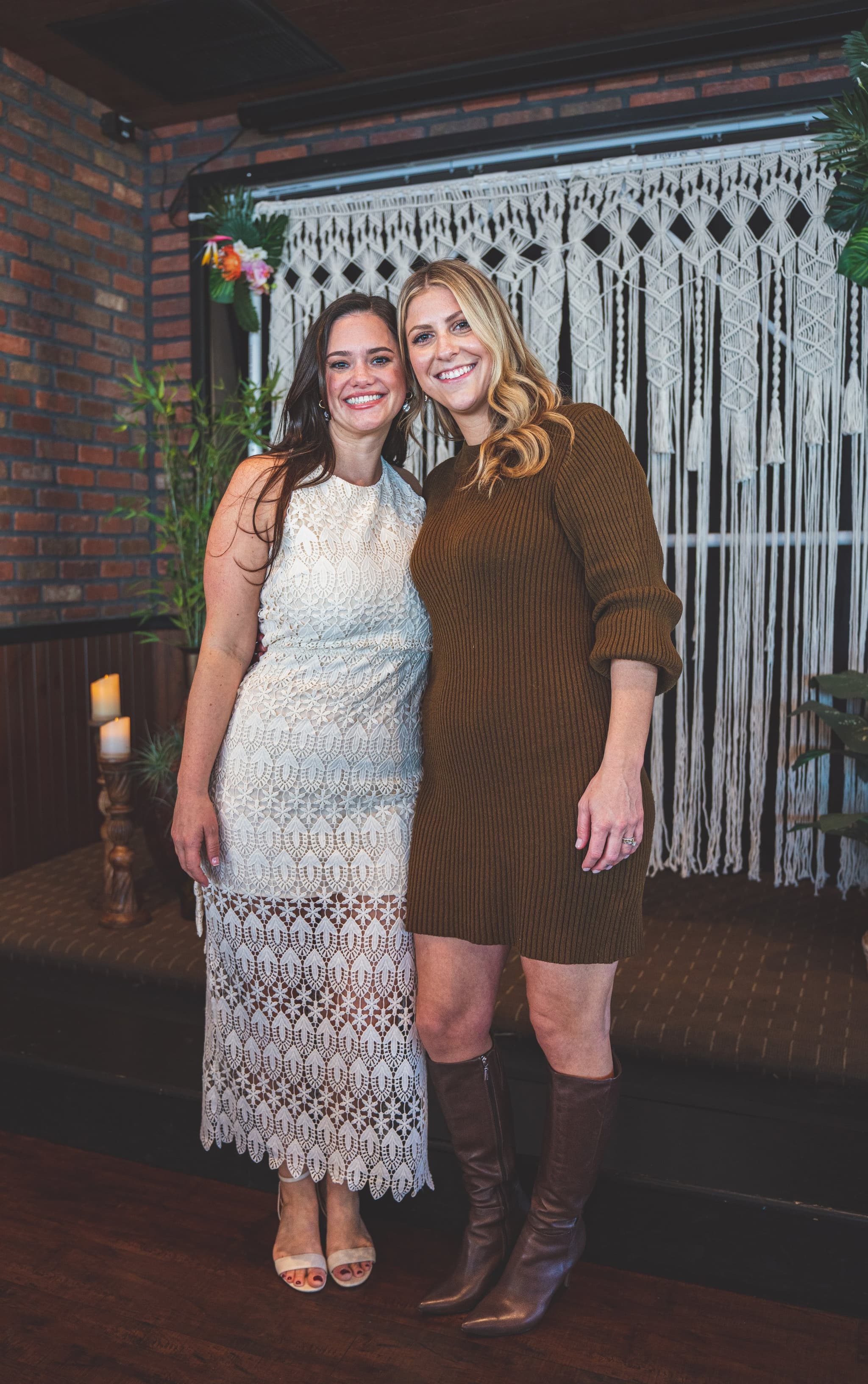 The bride in her white lace dress posing with two smiling friends in front of the boho macrame and tropical floral backdrop
