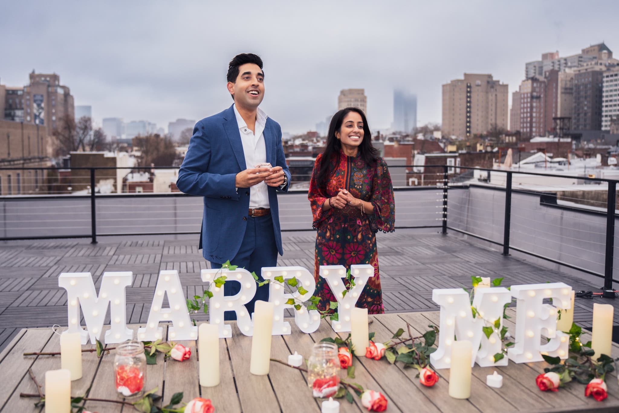 Couple laughing joyfully in front of lit MARRY ME sign with NYC skyline