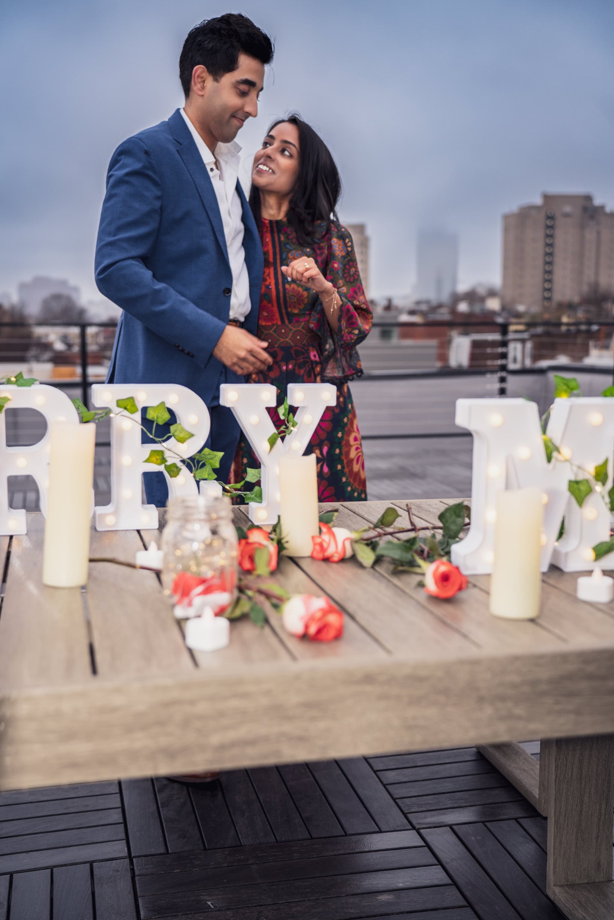 Couple laughing and leaning into each other in front of a romantic rooftop table decorated with candles, roses, and illuminated marquee letters