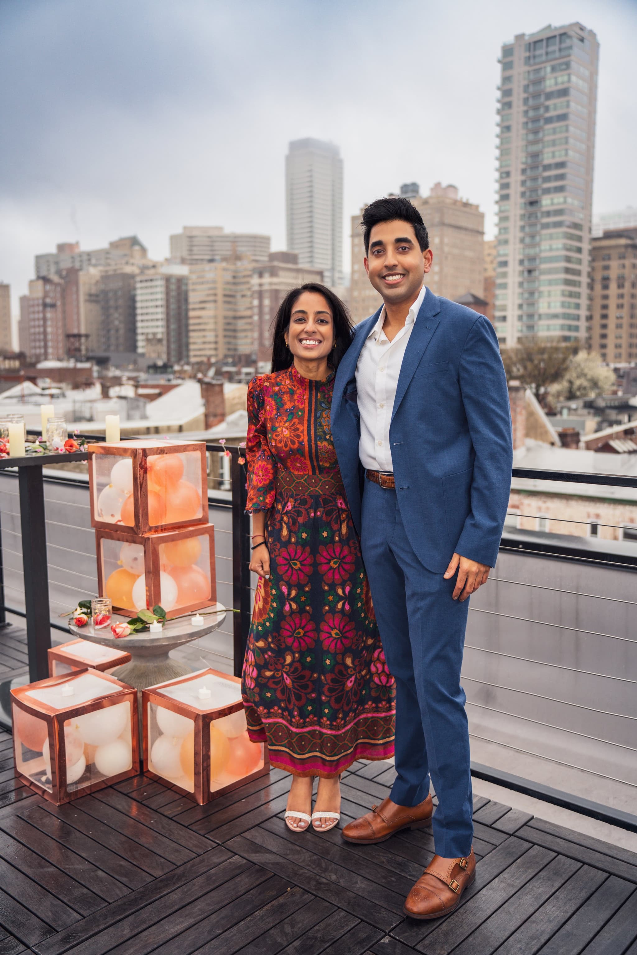 Couple posing together on a rooftop terrace with glowing balloon box decorations and the city skyline rising behind them