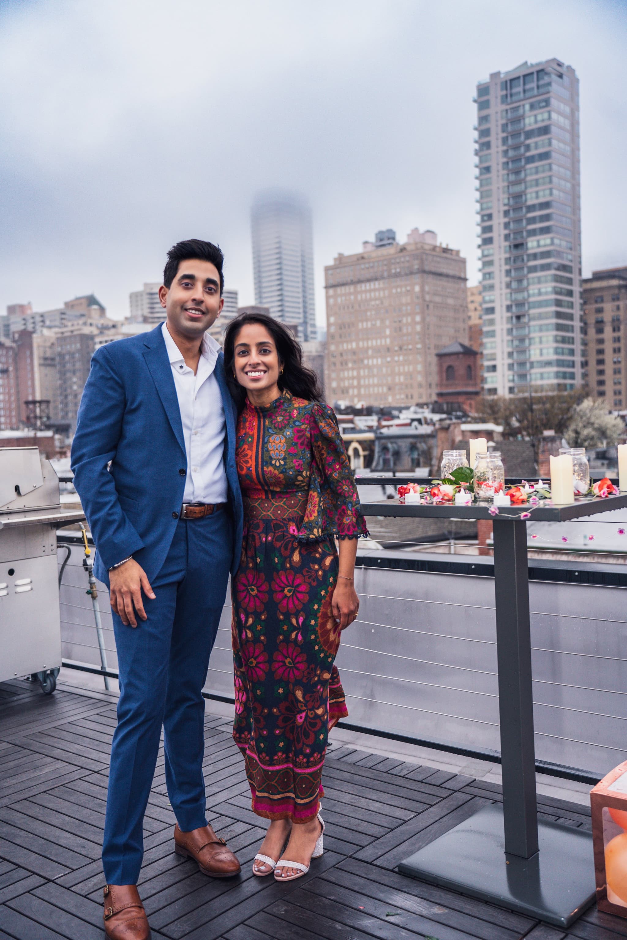 Newly engaged couple posing side by side on a rooftop terrace with the city skyline and candles and floral decorations beside them
