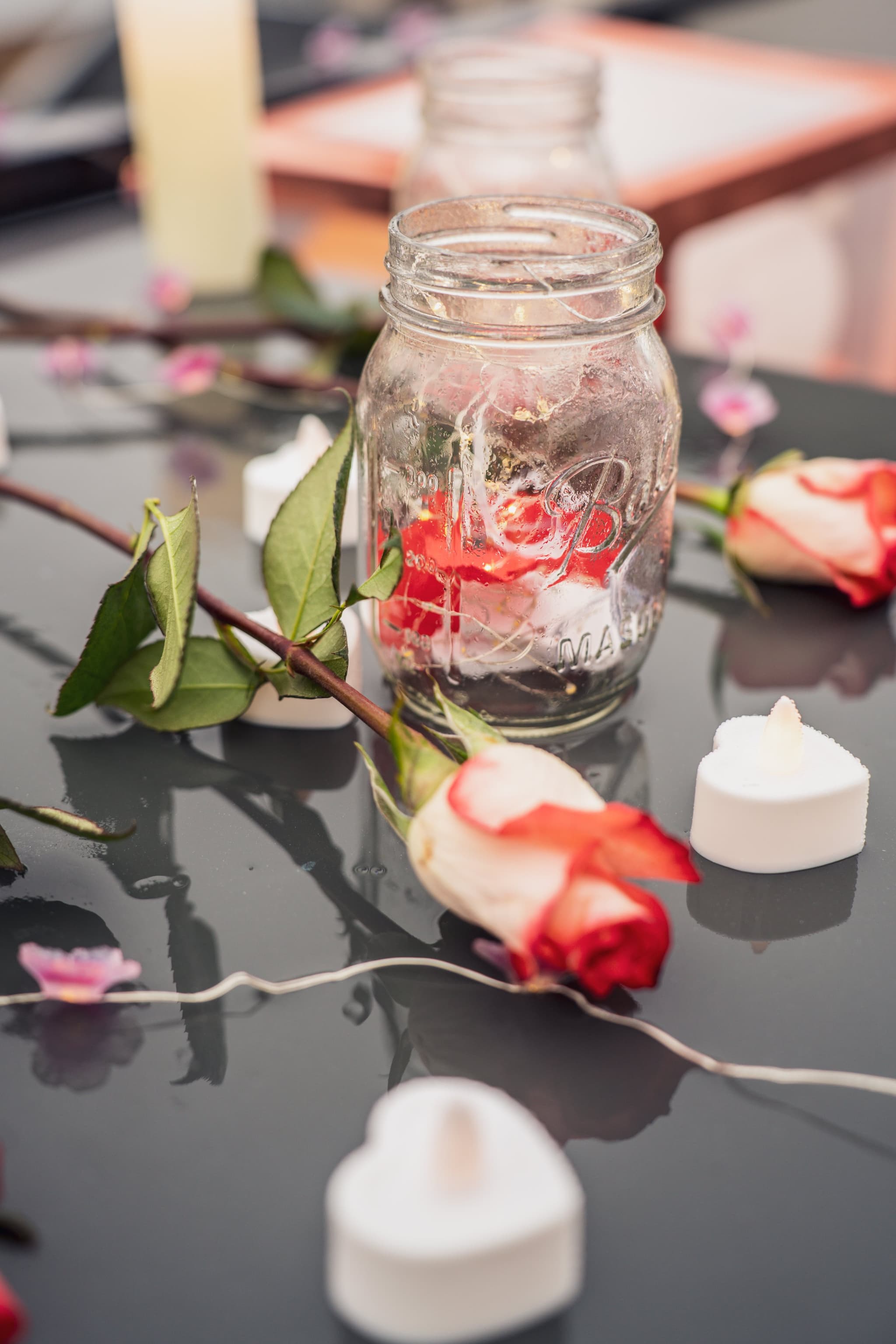 Romantic close-up of red and white roses scattered around a mason jar with candles glowing softly in the foreground