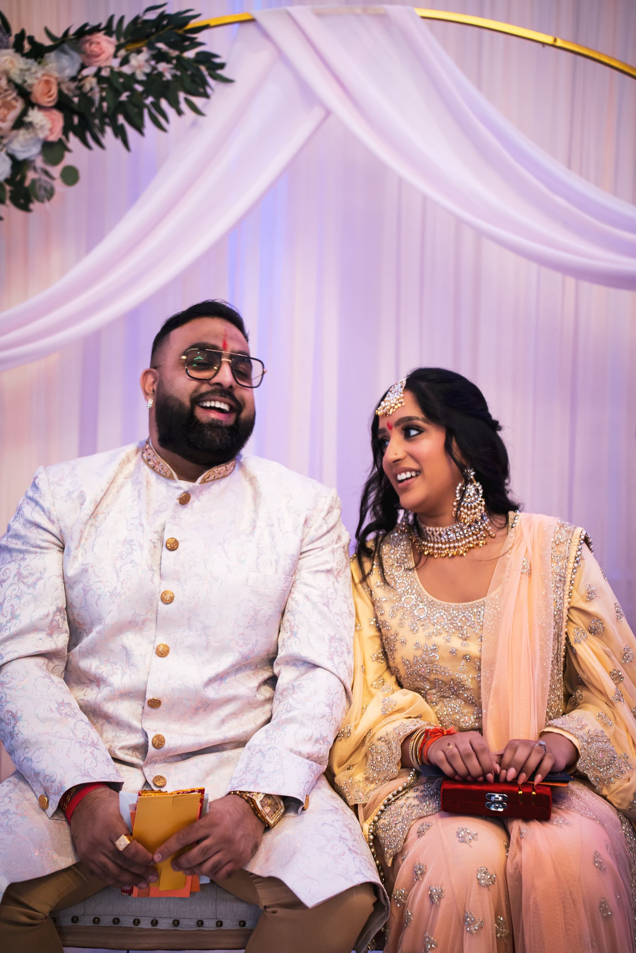 Couple shares a joyful candid laugh together while seated beneath a gold floral arch at their engagement celebration