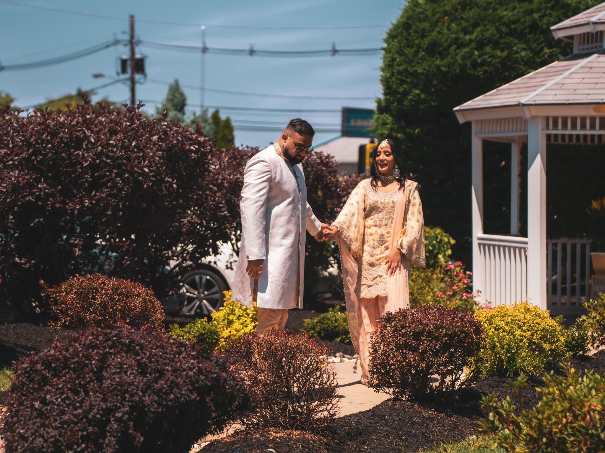Couple walks hand in hand through a lush garden path with a white gazebo and greenery as a sunlit outdoor backdrop