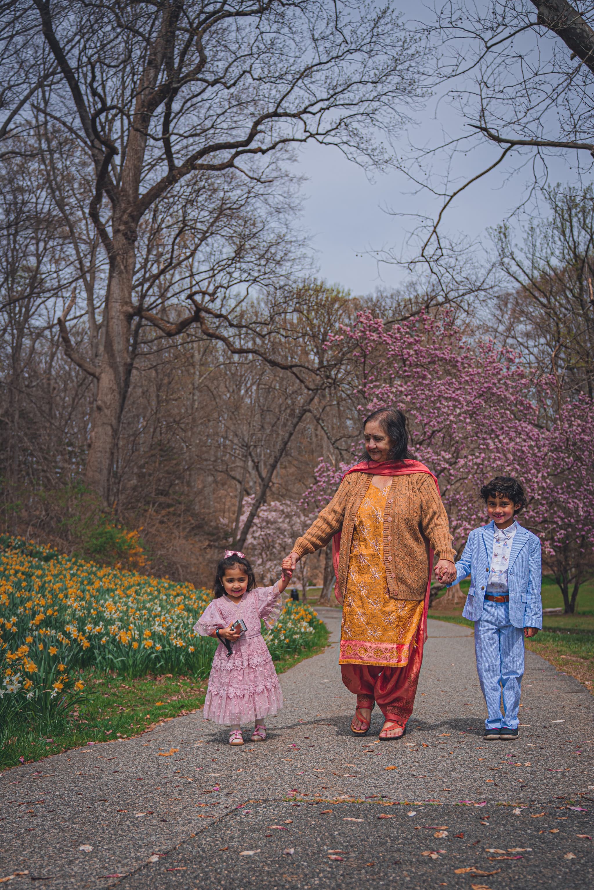 Mother walking hand-in-hand with two children along a park path lined with spring blossoms