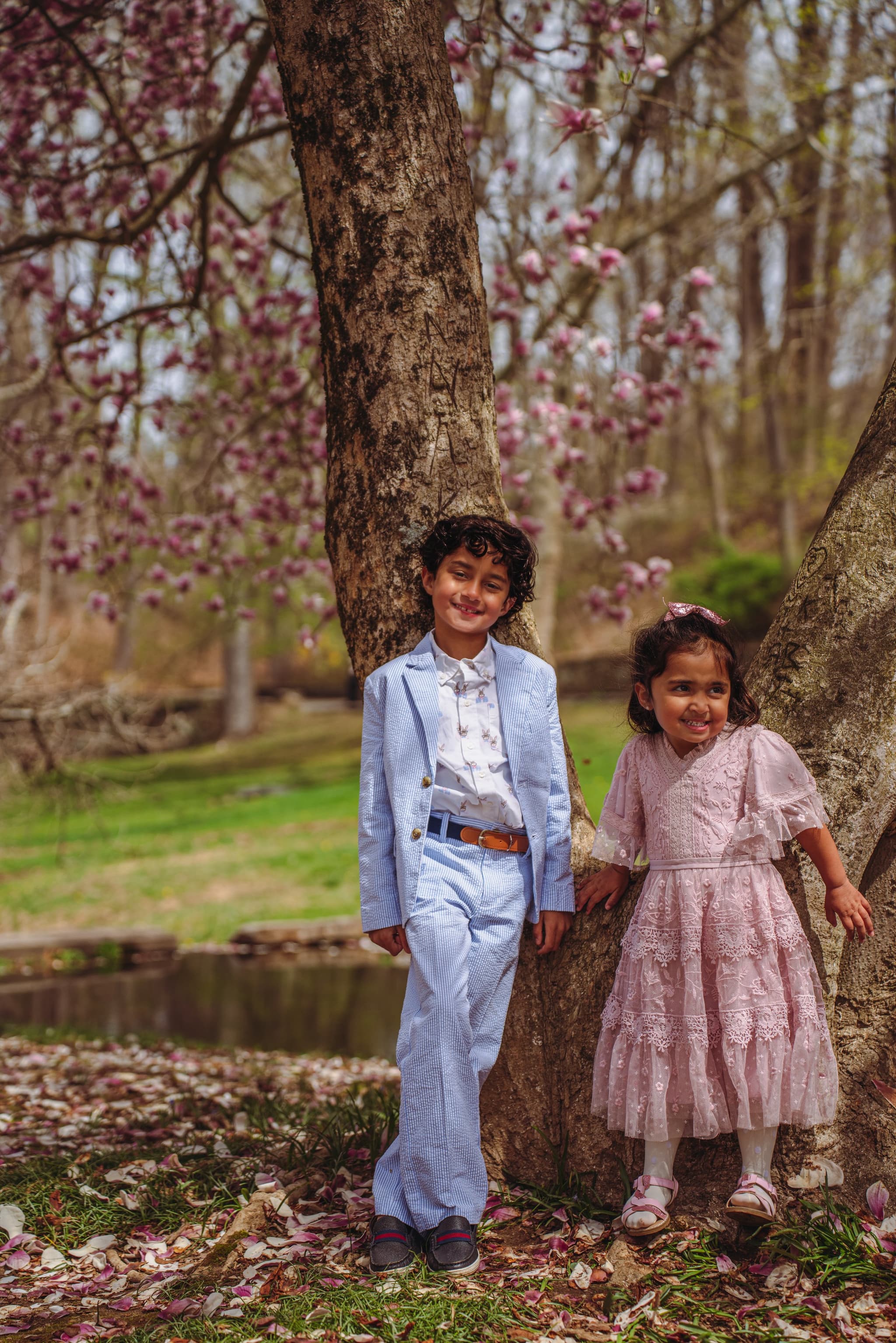 Siblings smiling and leaning against a tree trunk beneath pink spring blossoms