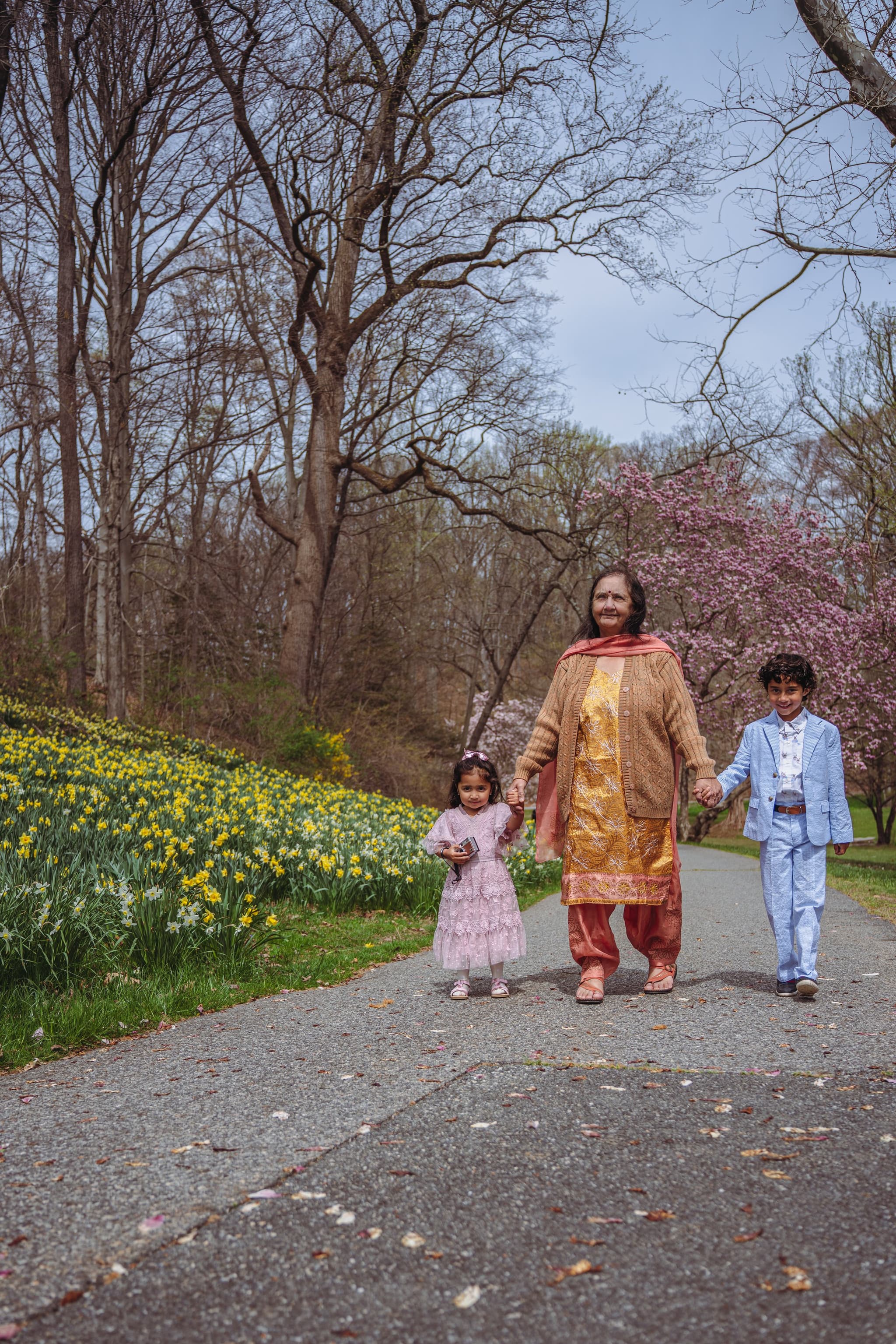 A mother walks hand-in-hand with her young son and daughter along a park path lined with daffodils and spring blossoms