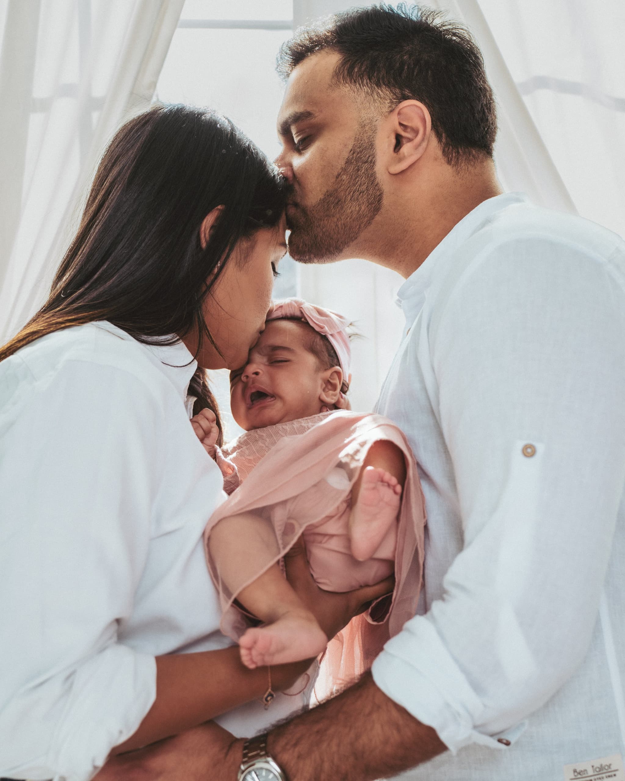 Parents leaning in to kiss their newborn, backlit by soft window light