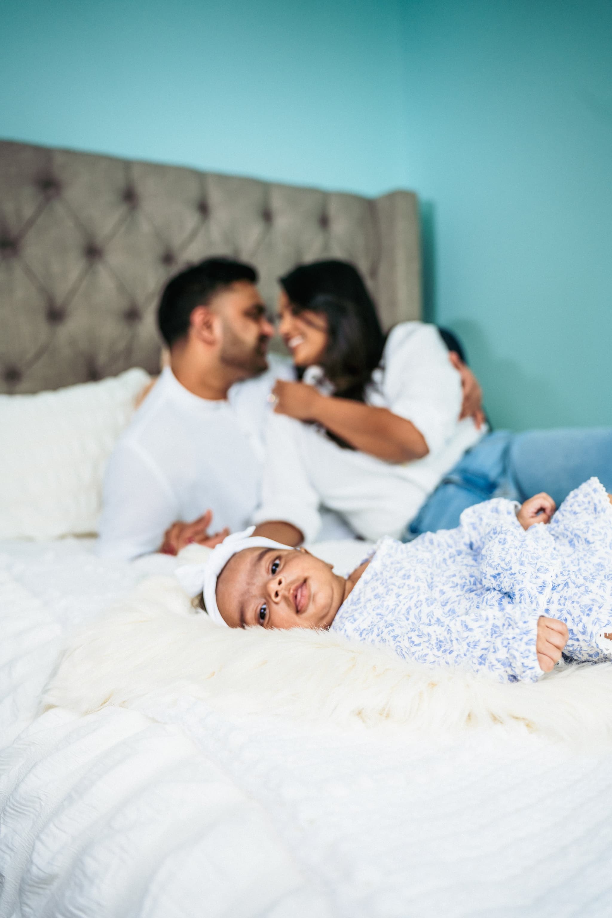 Baby alert in the foreground on white bedding, parents smiling softly in background