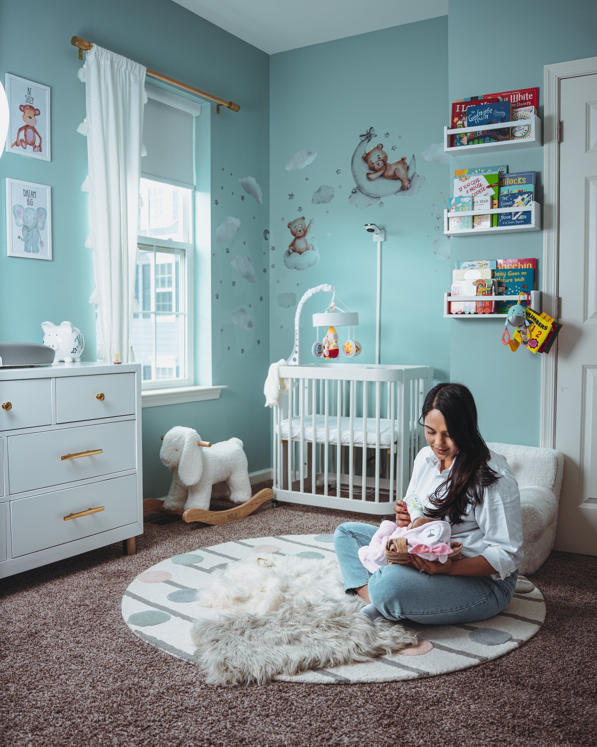 Mom sitting on nursery floor holding baby, full room with teal walls and crib visible