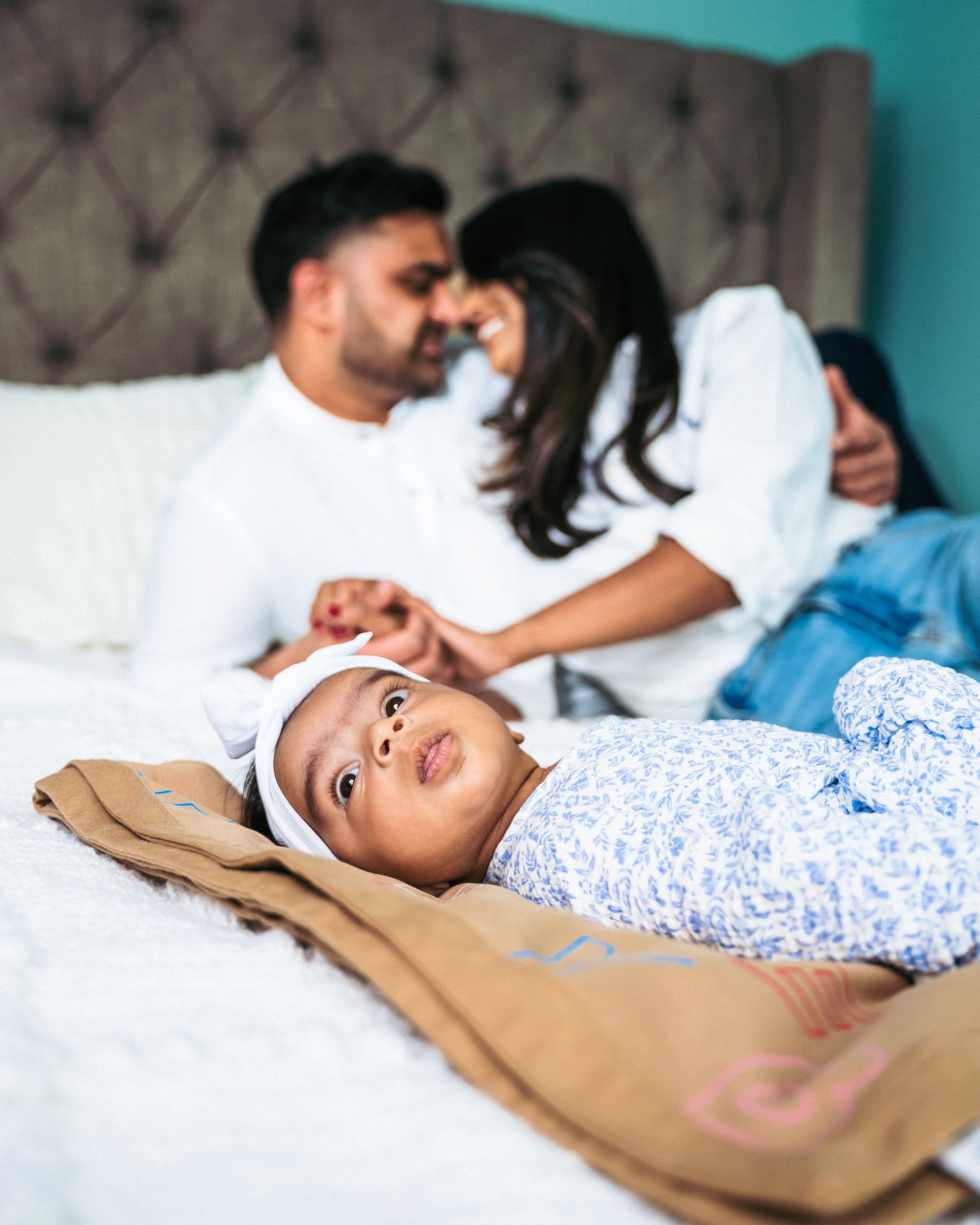 Newborn baby lying on the bed in the foreground while parents embrace lovingly in the background