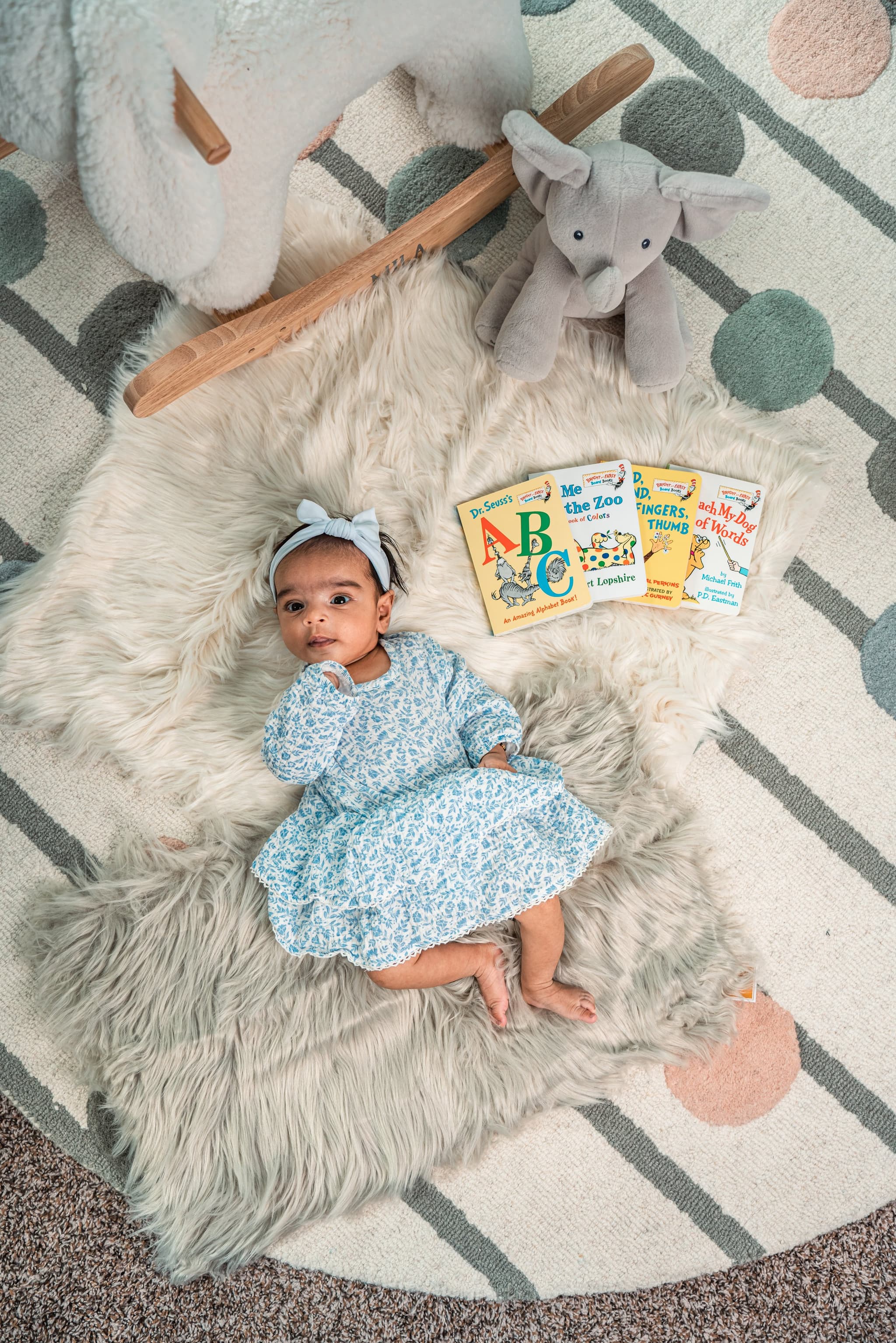 Newborn baby girl in blue floral dress lying on a fluffy rug surrounded by children's books and plush toys