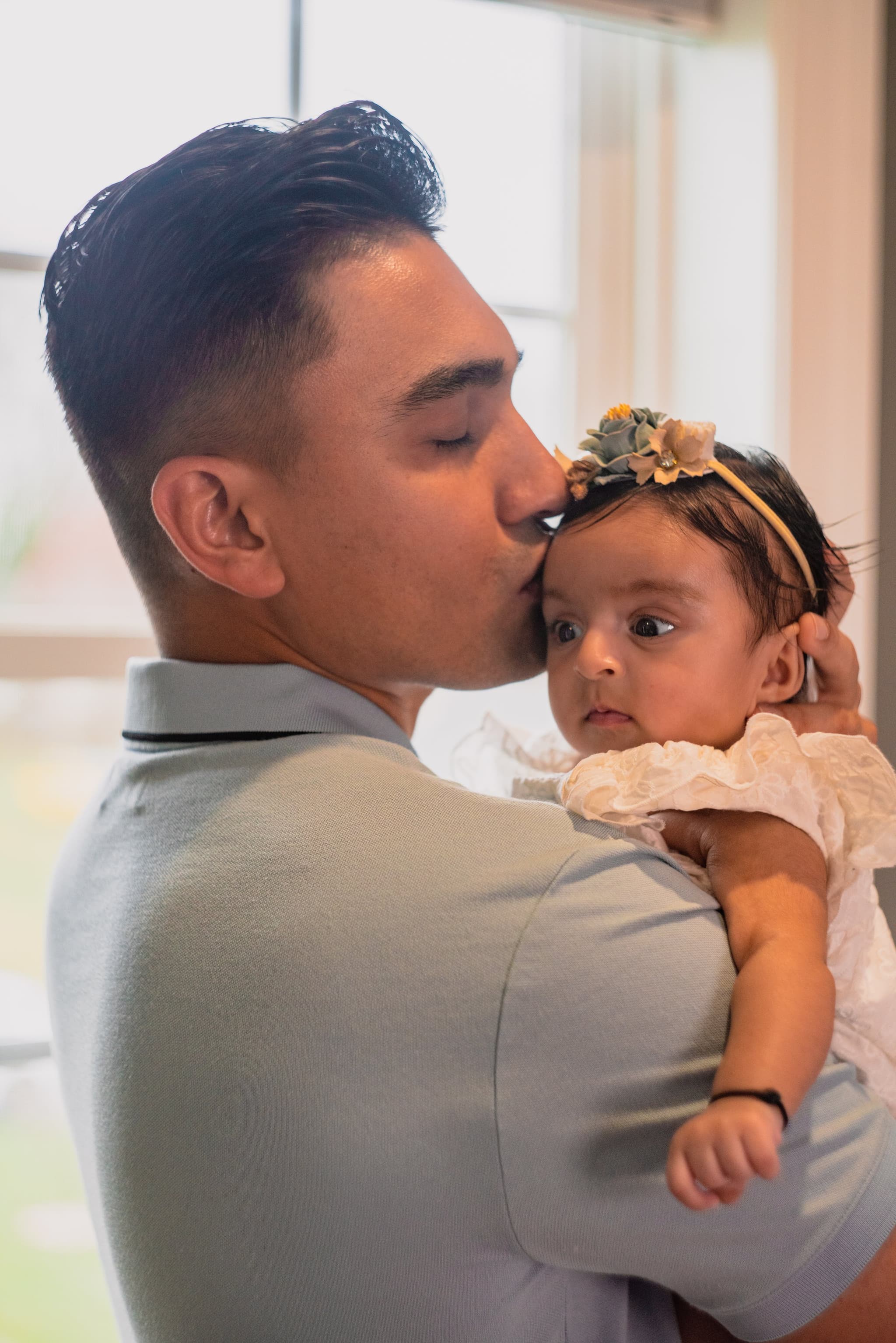 A father gently kisses his newborn baby's forehead while she looks up at him by a sunlit window