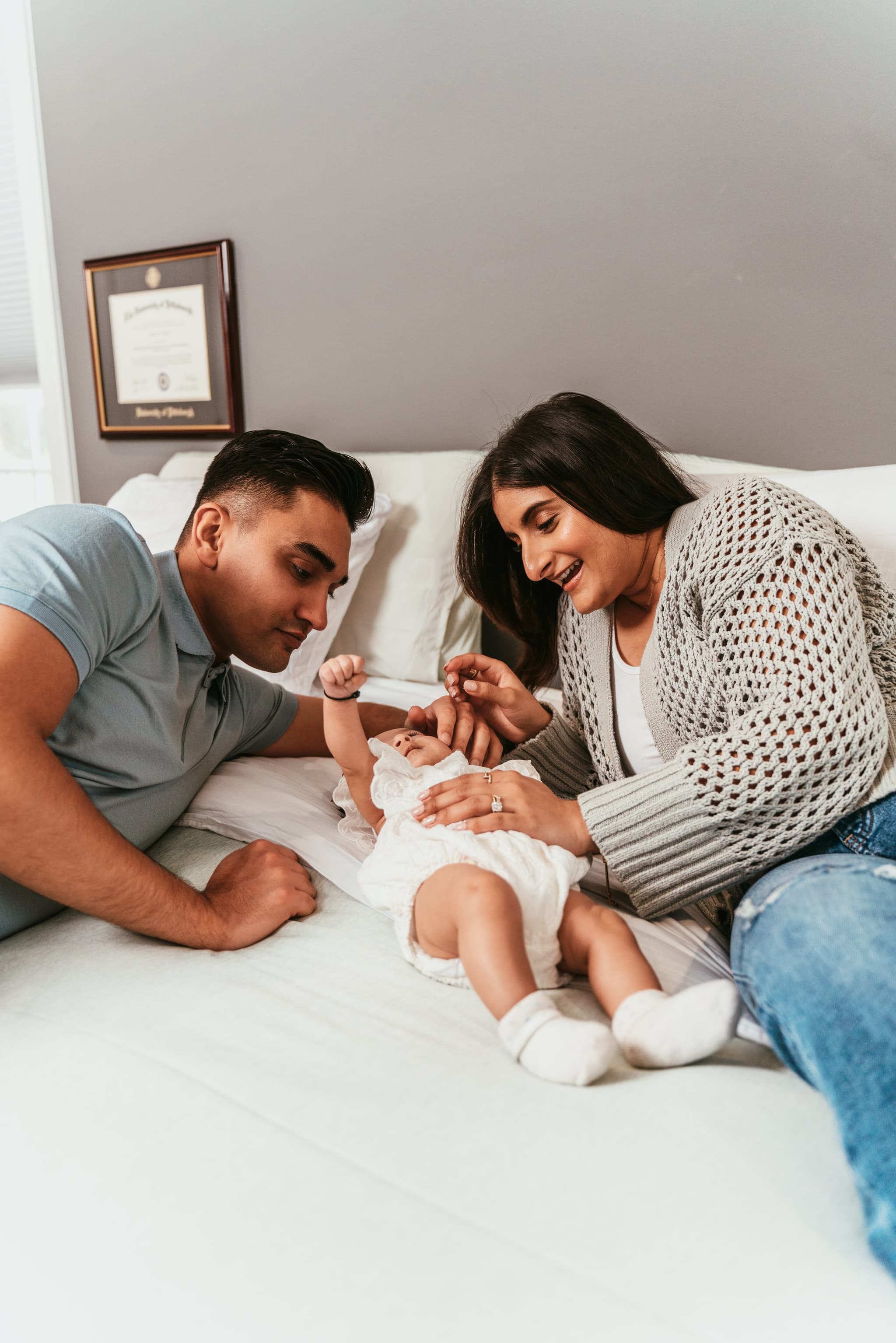 Both parents lie on a bed smiling and playing with their newborn baby girl between them