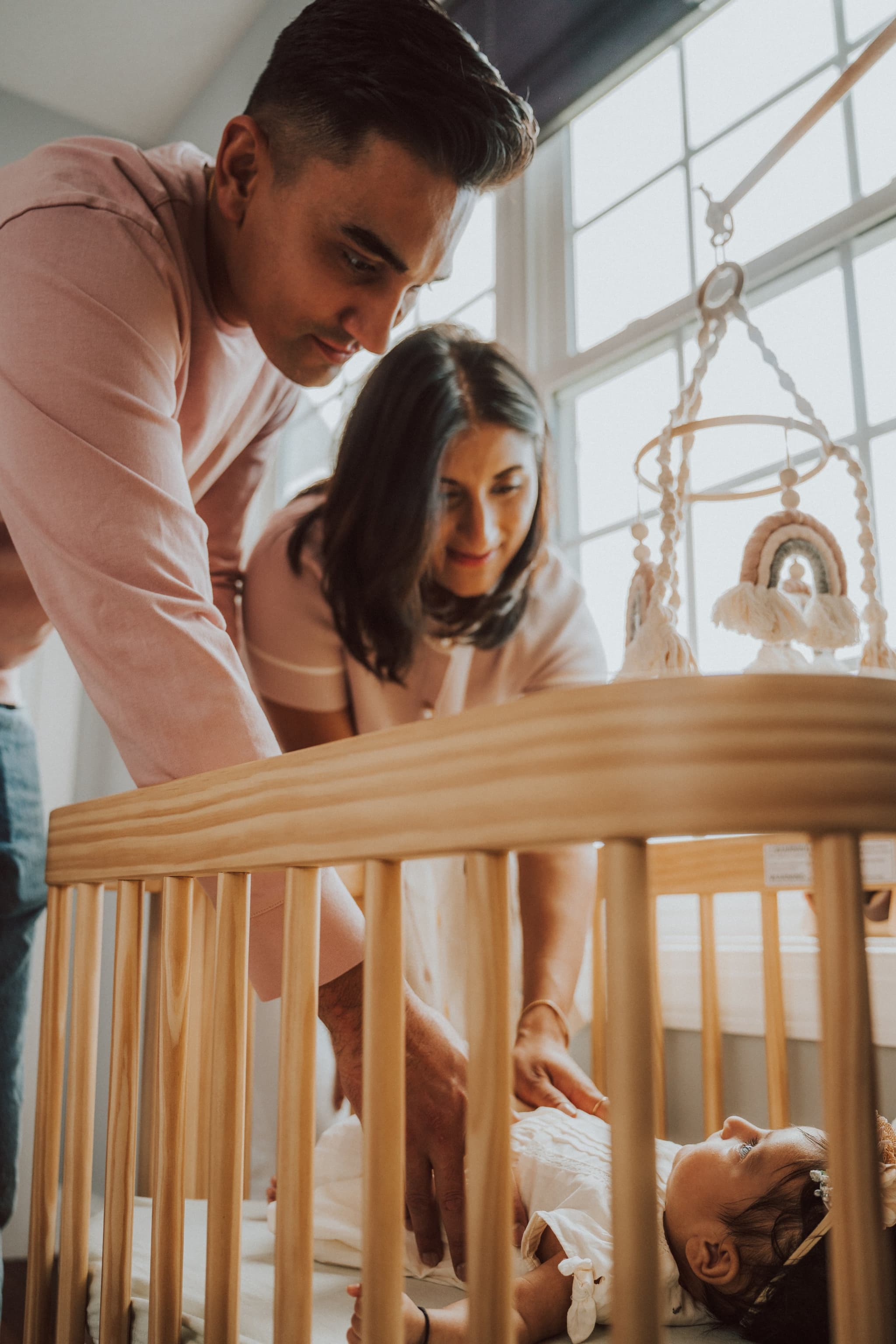 Parents lean over the crib together smiling at their newborn as soft window light fills the nursery