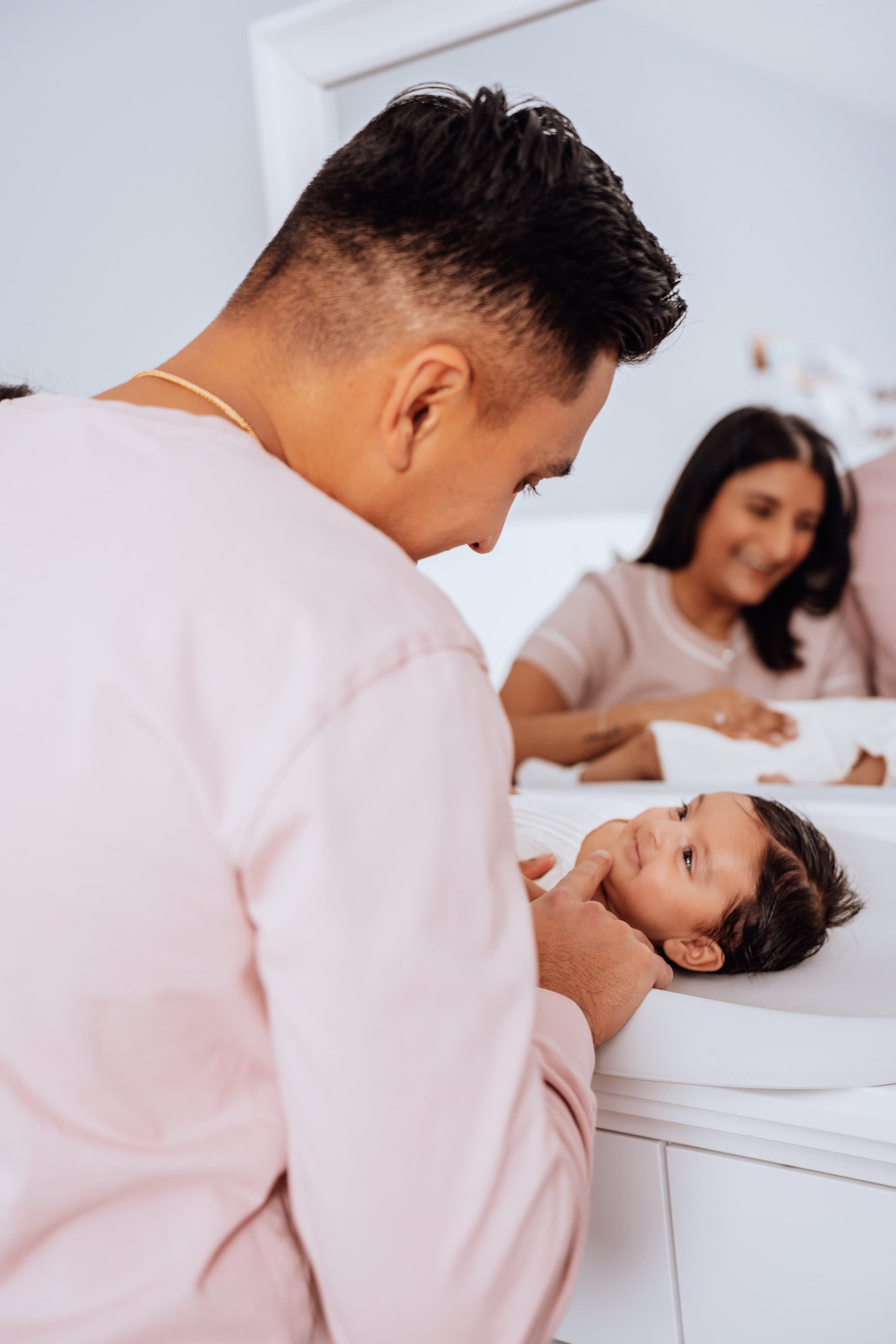 New family of three — mother, father, and newborn — snuggled together on a white bed in soft natural light