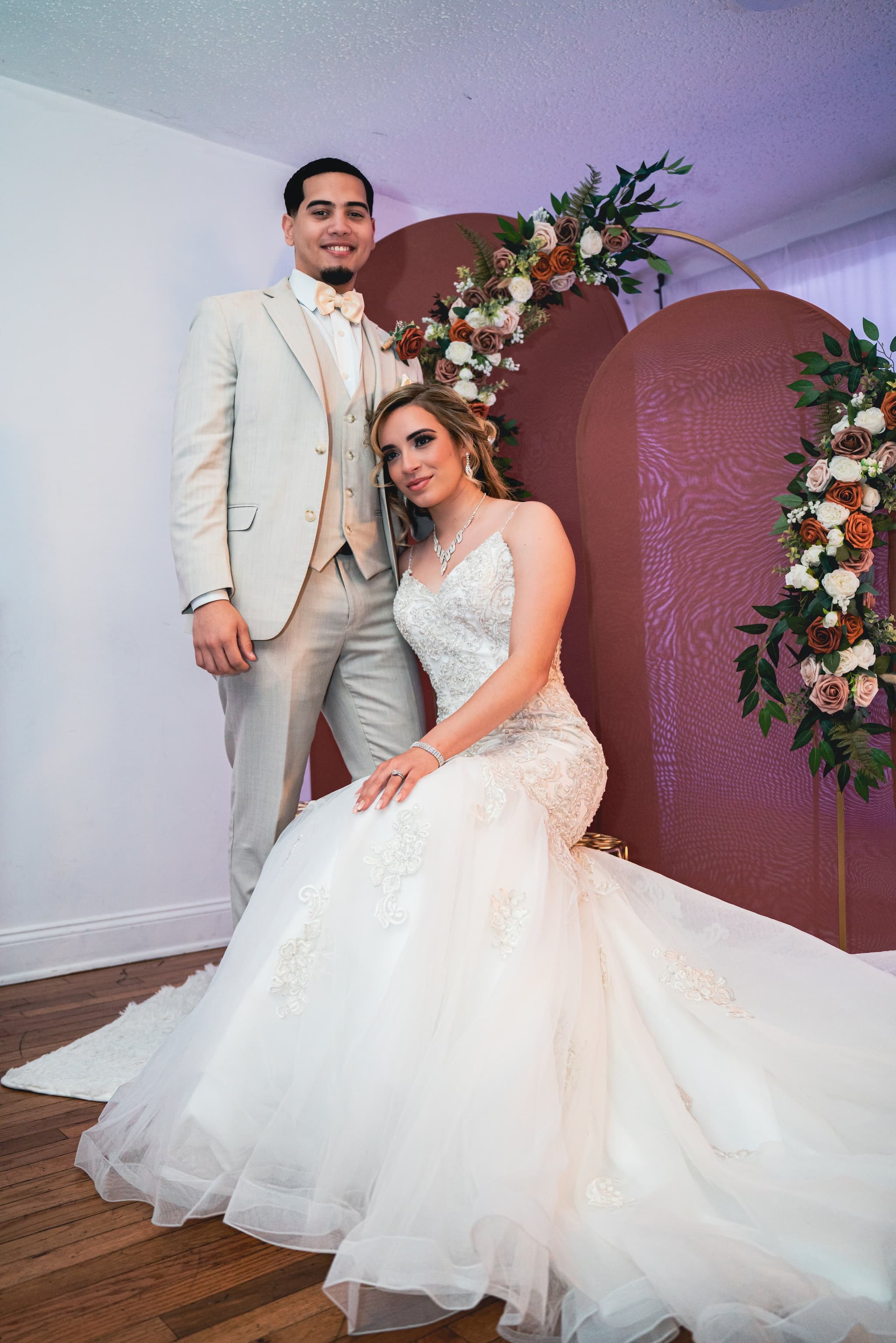 Couple posed in front of a terracotta floral arch, bride seated, warm tones
