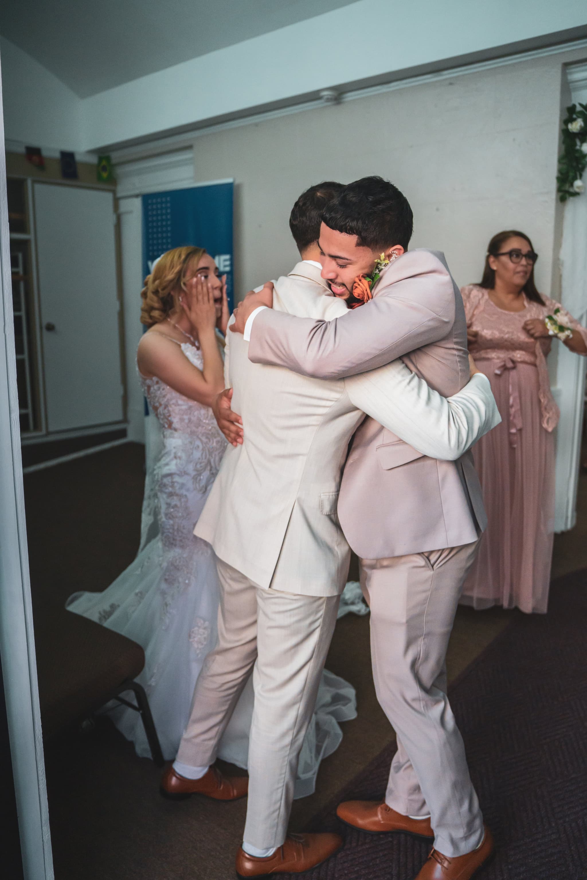 Candid emotional moment — groom embracing a close friend, bride reacting with hand over mouth