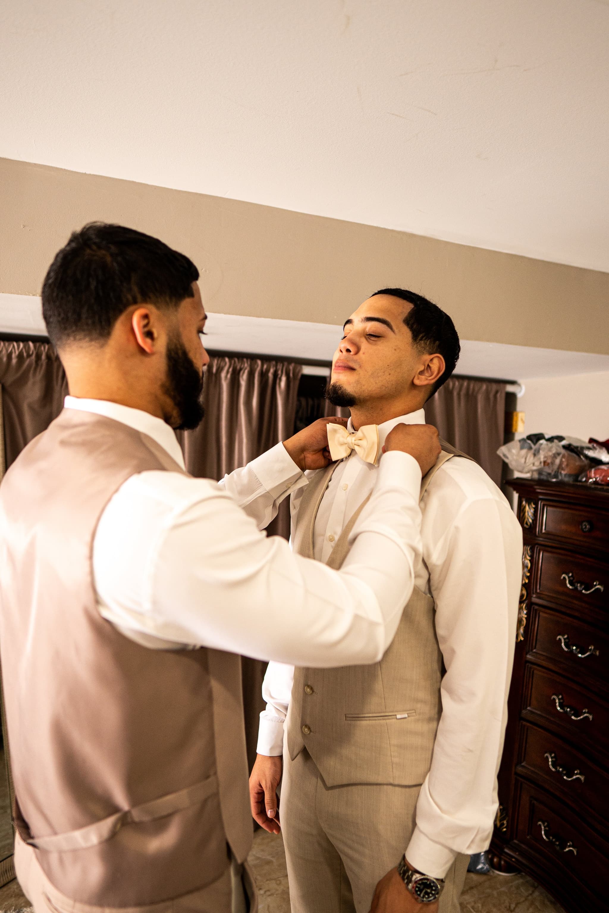 Groomsman adjusts the groom's bow tie while both share a calm moment during wedding preparations