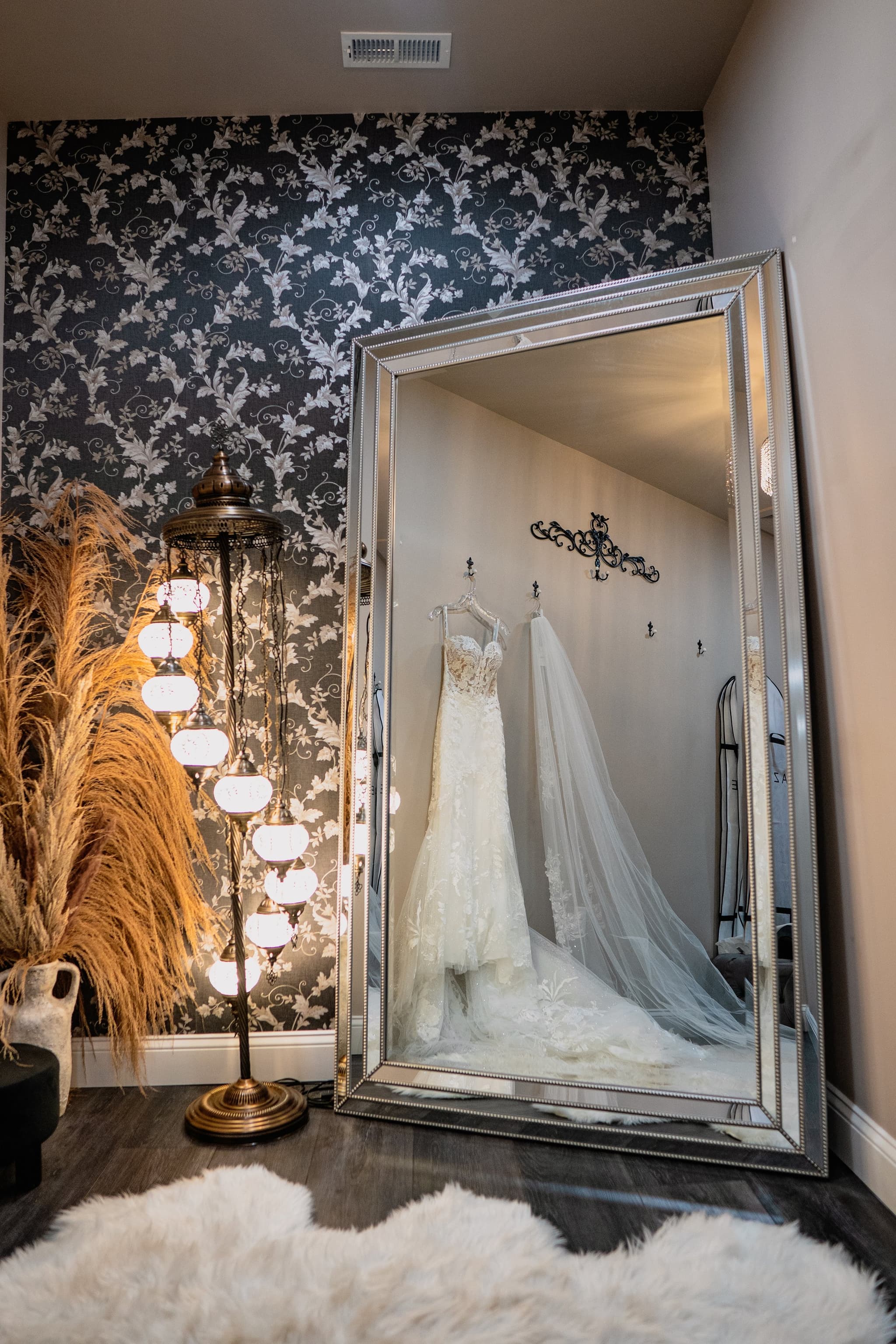 The bride's lace wedding gown and flowing veil displayed against a large ornate mirror surrounded by pampas grass and warm lamp light